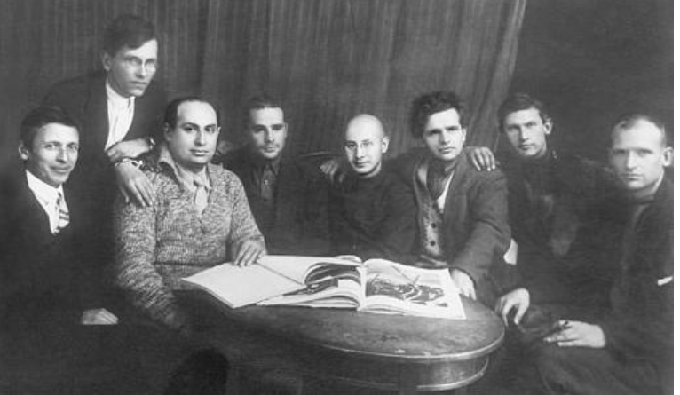 Eight men sit around a table with an open book, in a black-and-white photo, with a curtain backdrop, dressed in early 20th-century attire.