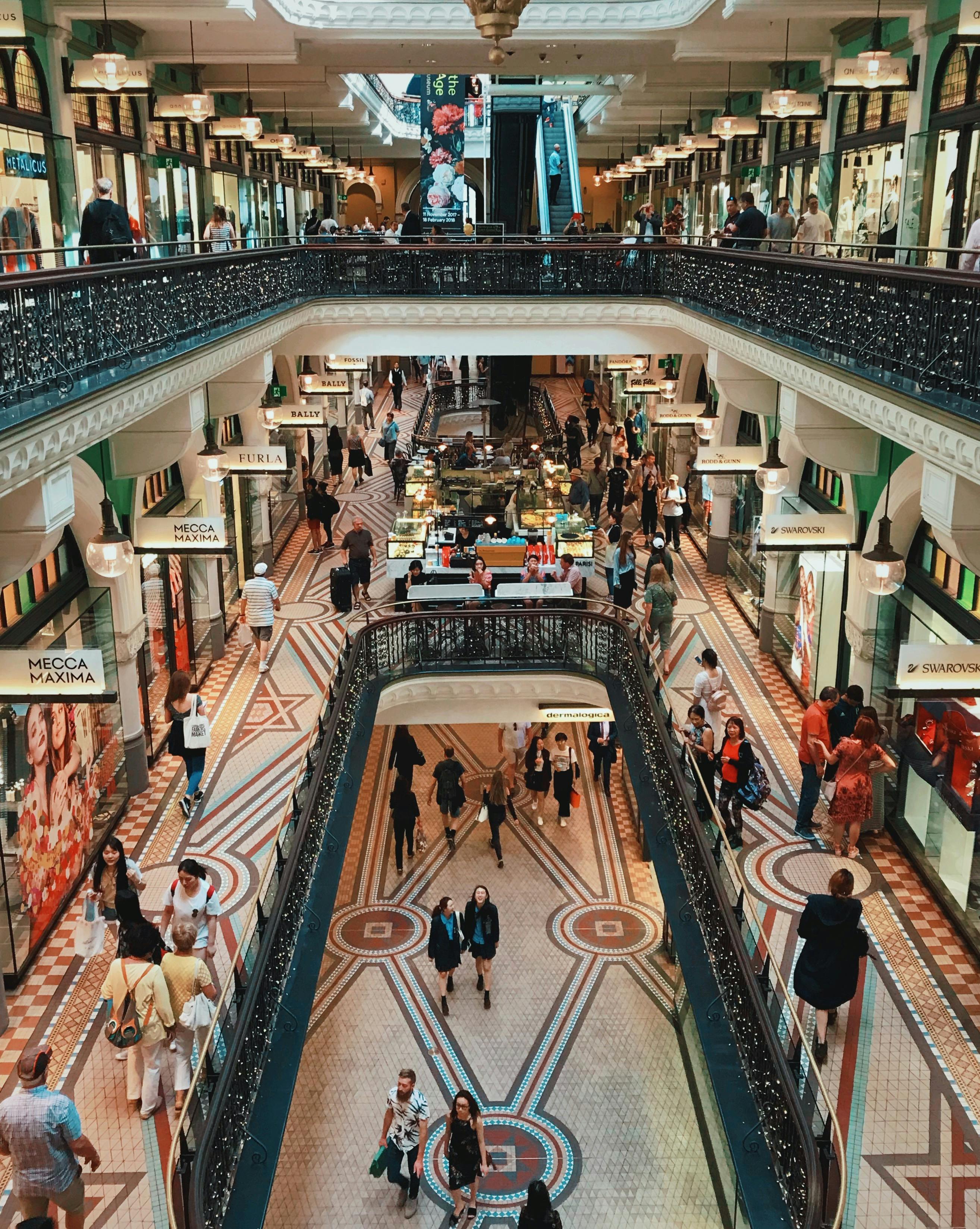 Busy multi-storey shopping centre interior with decorative iron railings, patterned tile floors, escalators, and numerous shoppers and storefronts.