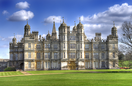 A grand historic mansion with ornate towers and architecture under a blue sky, surrounded by lush green lawns and a few trees.