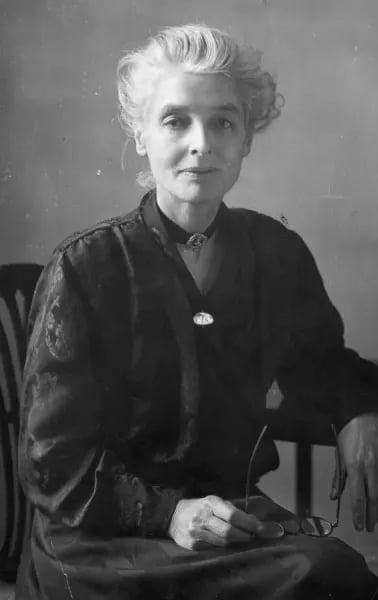 Elderly woman with short grey hair sits on a chair, wearing a dark dress and a necklace, holding glasses in one hand against a plain backdrop.