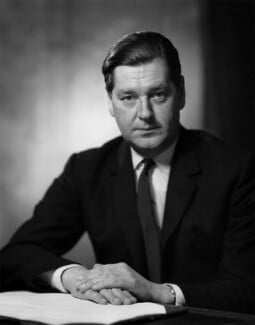 Man in a suit sits at a table with hands clasped, looking forward. The background is blurred and he has neatly combed hair, exuding formality.