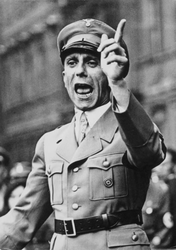 A man in a military uniform is passionately speaking, with his right arm raised and finger pointing, in a historical black and white photo.