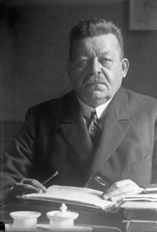 Elderly man with moustache in formal suit seated at a desk with papers and pens, looking directly at the camera in a dimly lit setting.