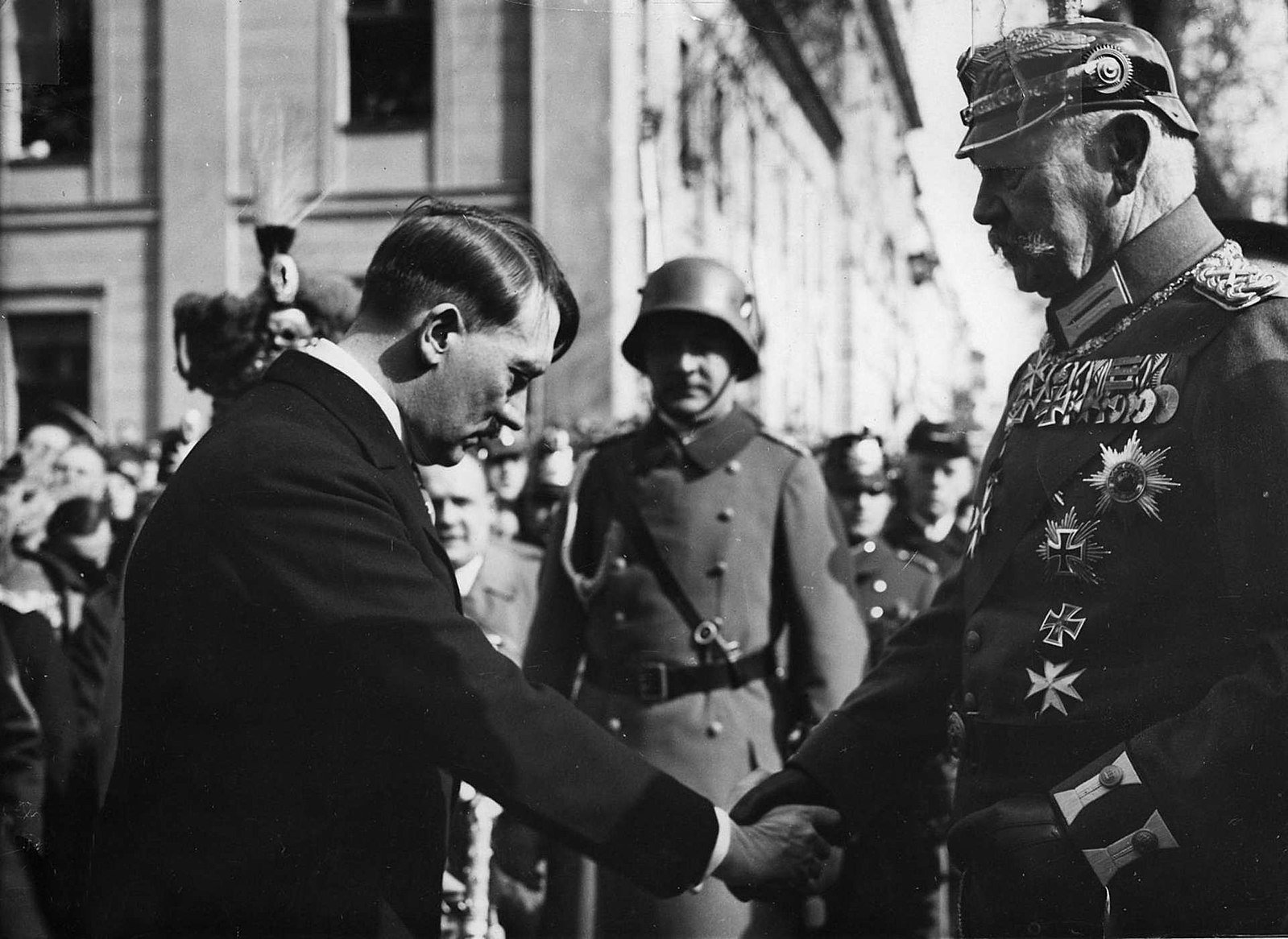 A man in a suit shakes hands with a uniformed man wearing numerous medals, surrounded by other uniformed individuals in a formal setting.