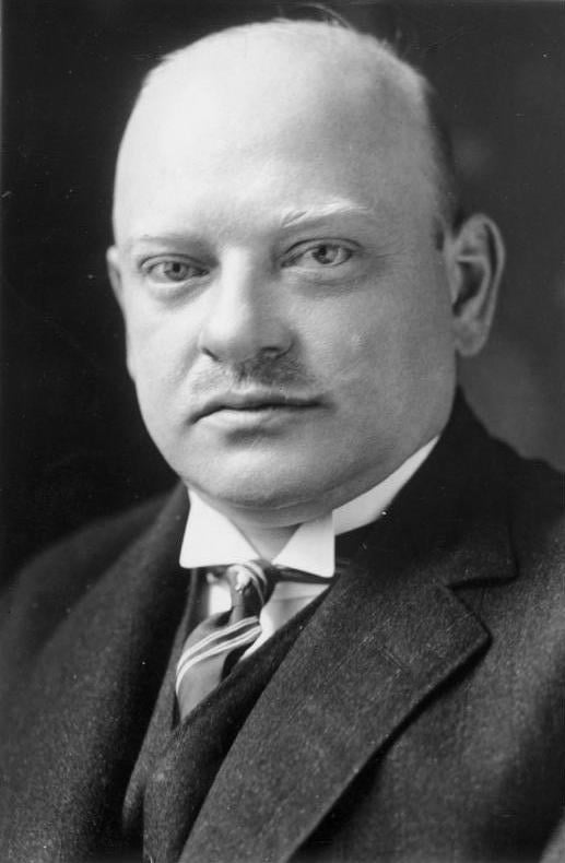 Bald man in an early 20th-century suit and tie, with a stern expression, facing forward against a dark background, likely a formal portrait.