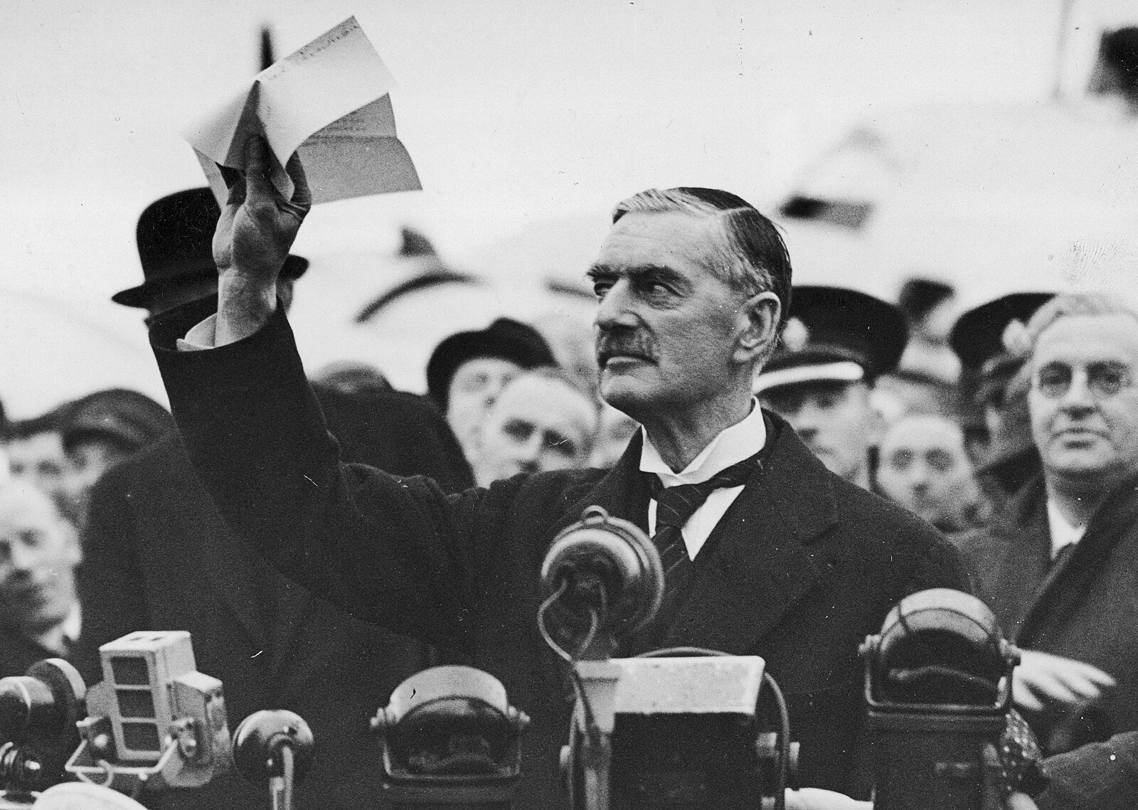 A man in a suit holds up a paper amid a crowd, speaking into multiple microphones, suggesting a historical outdoor event or announcement.