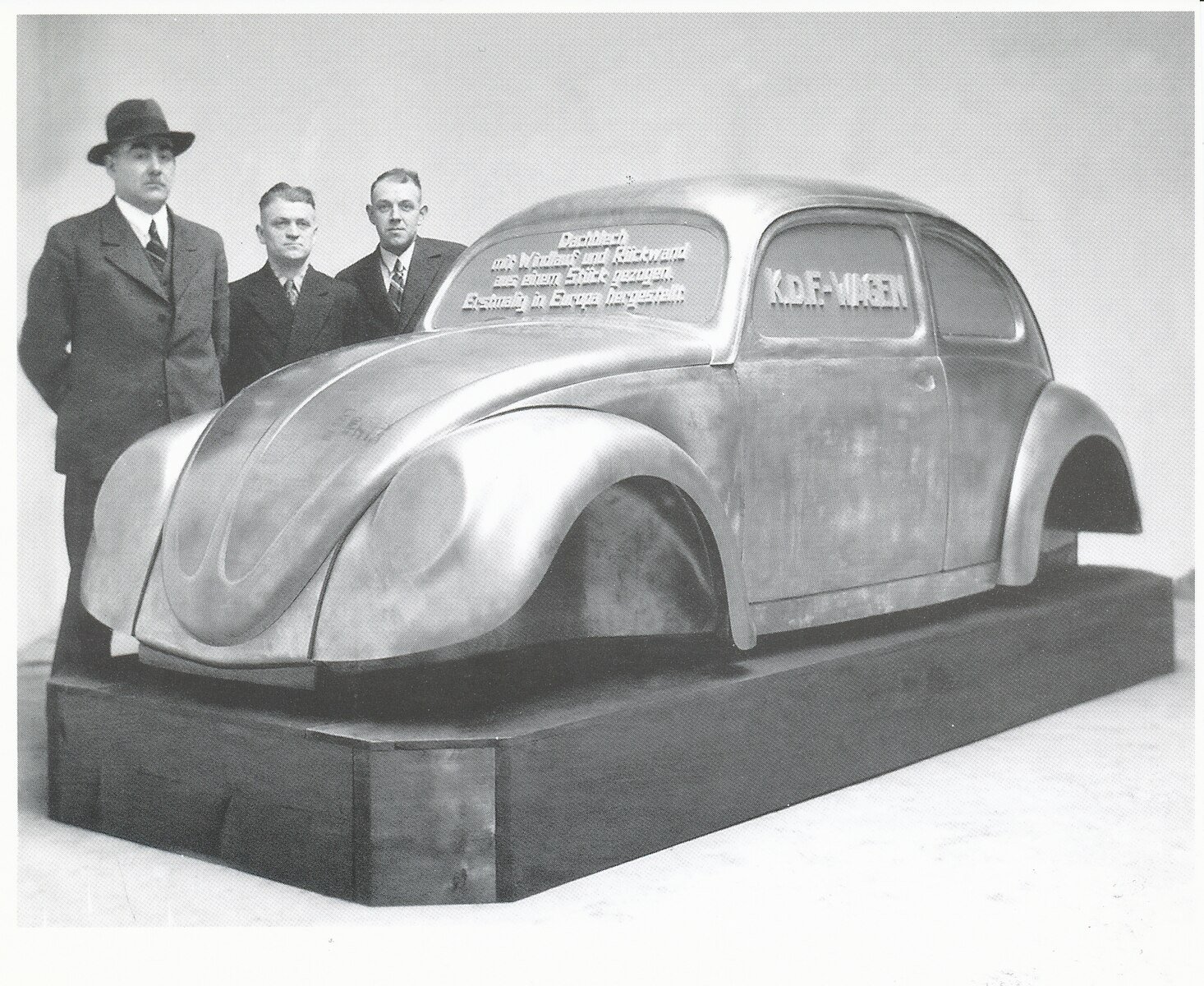 Three men stand beside a metal prototype of a car marked "KdF-Wagen," showcasing a streamlined design, on a display platform in a studio setting.