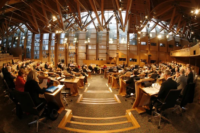 Wide view of a parliamentary chamber with delegates seated in curved rows, wooden interior, and large windows with natural light streaming in.