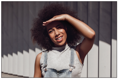 Person with curly hair wearing a grey top and denim overalls, smiling and shading their eyes from the sun with one hand, against a striped wall.