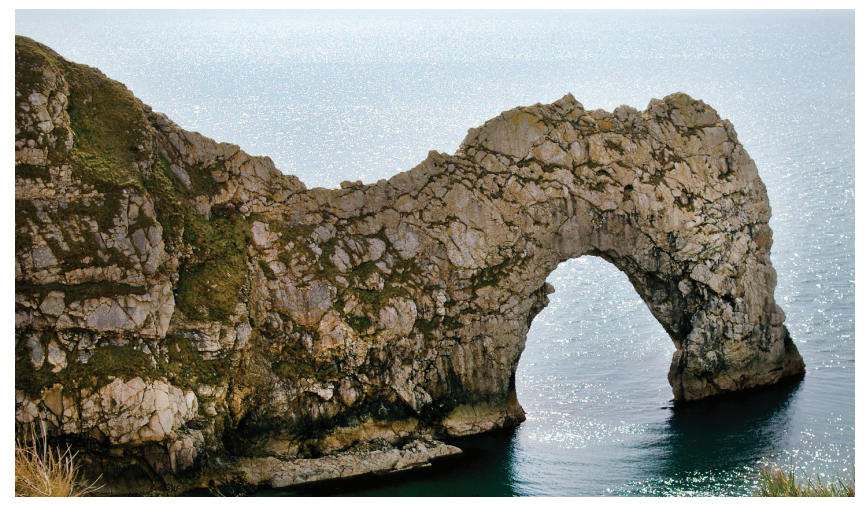 Natural stone arch with rugged cliffs, known as Durdle Door, extends into a calm sea under a bright sky, with sunlight shimmering on the water.