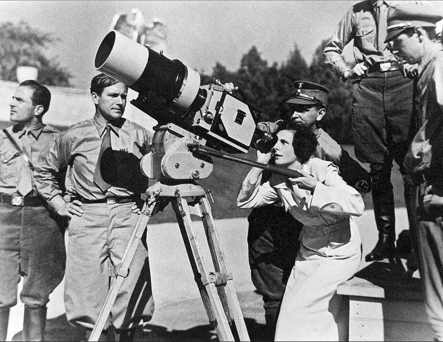 A group of men in uniform observe a woman using a large camera on a tripod outdoors, set against a blurred background of trees and buildings.