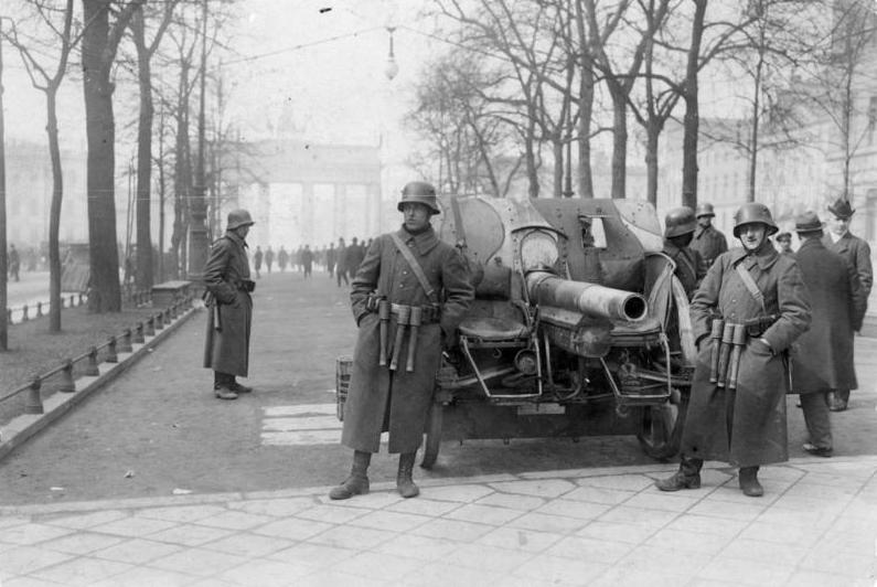 Soldiers in long coats and helmets stand near a large artillery gun on a city street, with rows of trees and a prominent gate in the background.