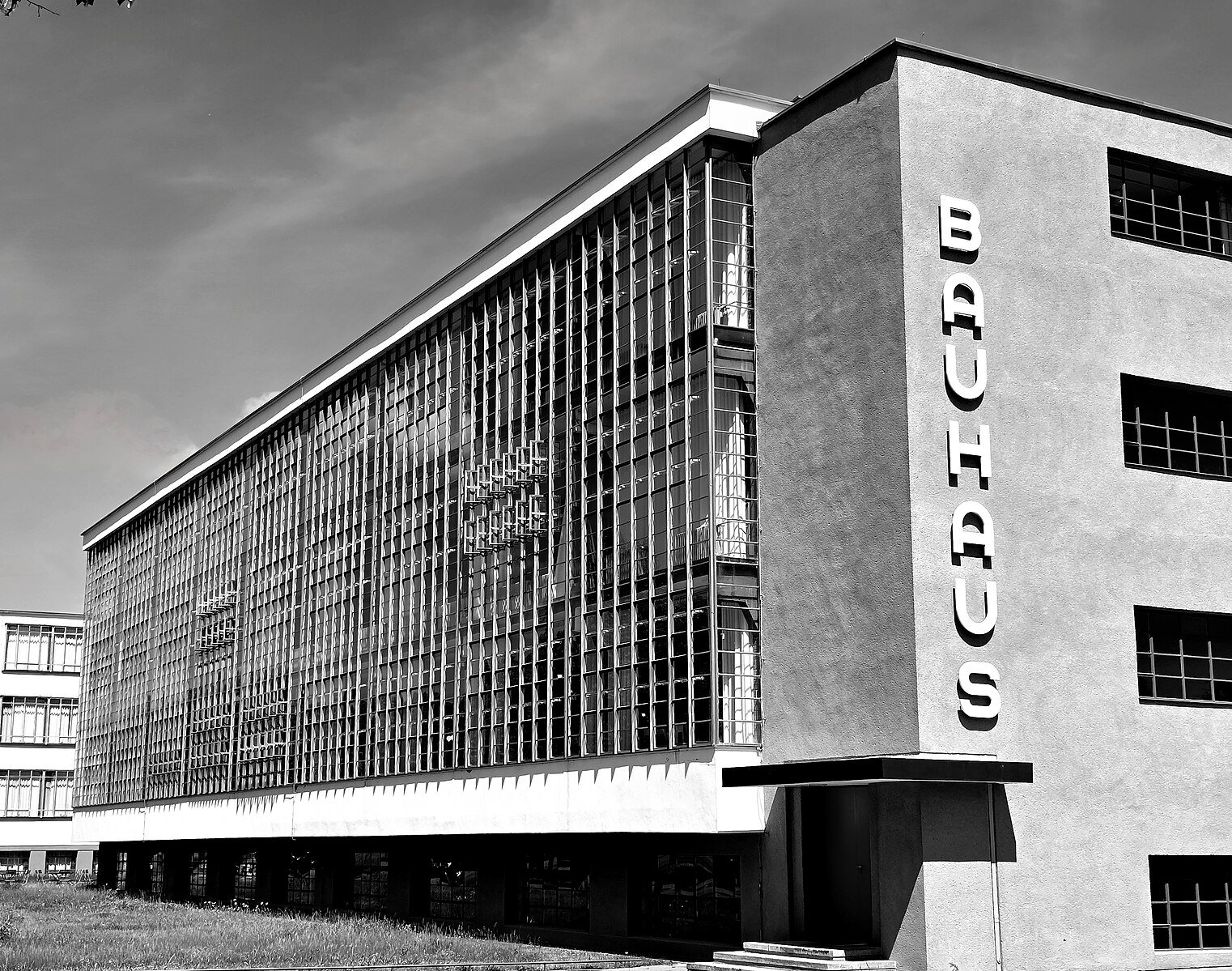 Black and white image of Bauhaus building with vertical glass windows, flat roof, and "BAUHAUS" text on the side; grass lawn in foreground.