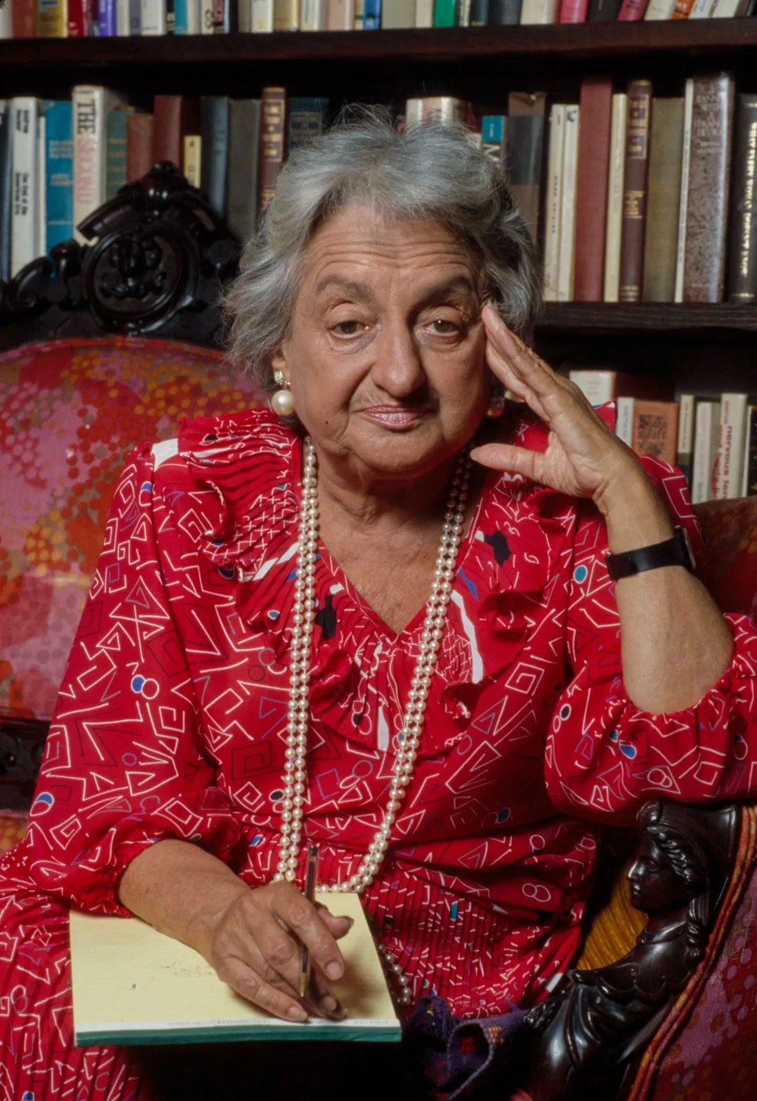 Elderly woman in a red patterned dress, seated with a notebook and pen, wears pearl necklace, rests her head on hand, bookshelf behind her.