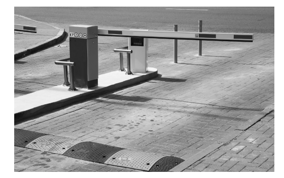 Car park barrier gate arm closed, flanked by two bollards on a brick-paved driveway. Speed bump visible in the foreground.
