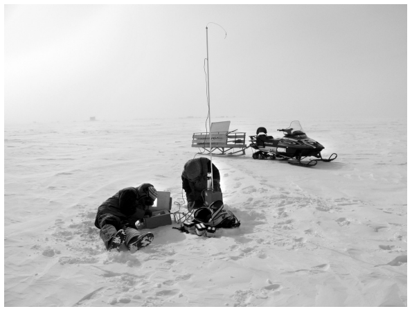 Two people work on equipment in a snowy field; a snowmobile and sled are nearby, suggesting a remote, cold environment for scientific research.