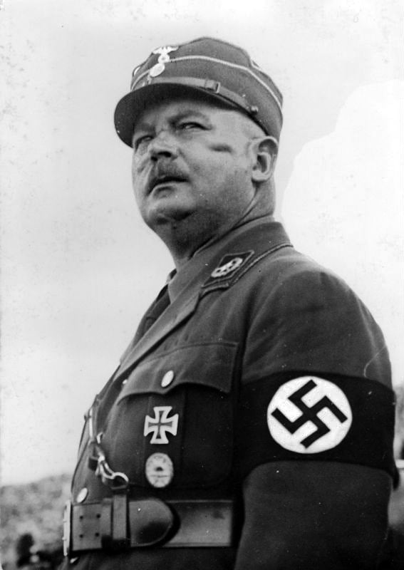 A man in a military uniform with insignias and an armband, looking upwards. The background is a cloudy sky.