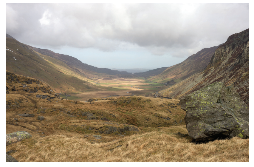 Mountain valley landscape with rocky foreground and grassy hills, cloudy sky above, and distant fields in the background.