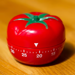 Red kitchen timer shaped like a tomato with green leaves, displaying numbers 5 to 30 around its middle, set on a wooden surface.