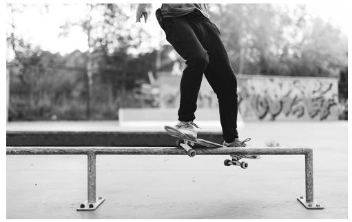 Person skateboards on a rail in a park, performing a trick. Background features trees and graffiti on a wall, captured in black and white.