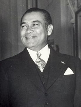 Smiling man in a suit and tie with a pocket square, standing in a formal setting, facing slightly left, with a blurred doorway in the background.