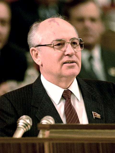 Mikhail Gorbachev is shown in the picture. An elderly man in a suit with glasses, speaking into microphones at a conference, faces the audience with blurred people in the background.
