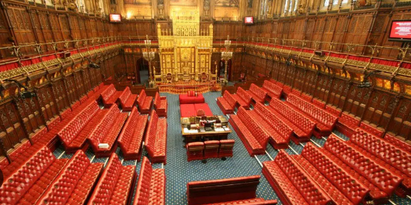 Interior of the House of Lords, UK Parliament, featuring red leather benches, ornate wood panelling, and a gold throne-like structure at the far end.