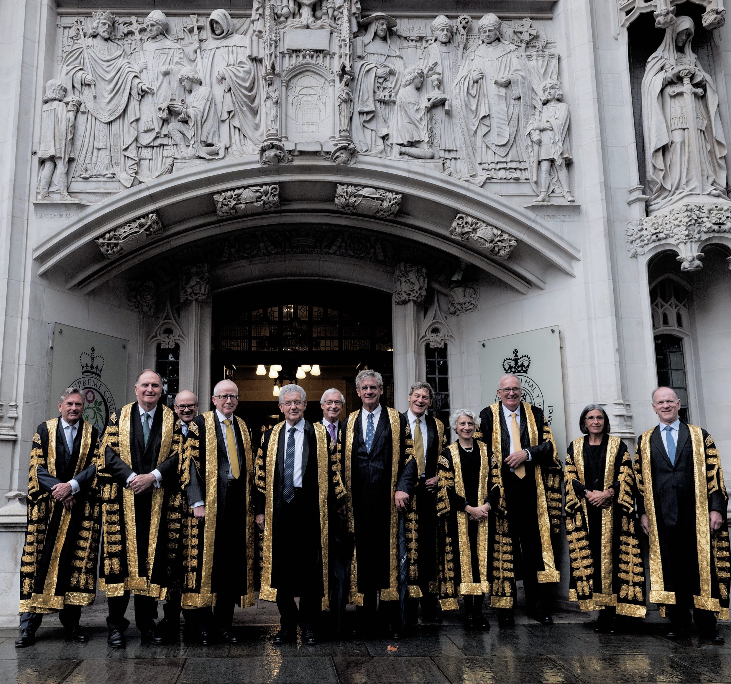 A group of judges in ornate robes stand in front of a building with intricate stone carvings, possibly the UK Supreme Court entrance.