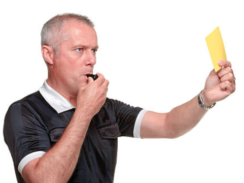 Referee in a black uniform blowing a whistle, holding a yellow card up against a plain white background.