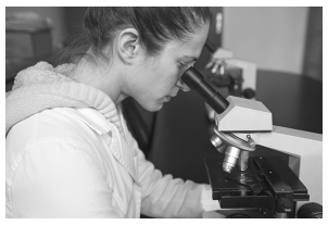 A woman in a lab coat uses a microscope in a laboratory setting, focusing attentively on a sample slide under the lens.