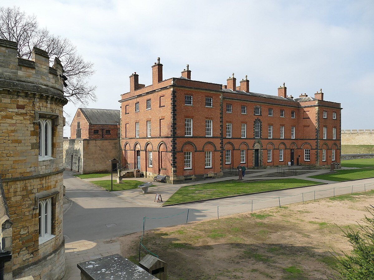 Historic brick building with arched windows, surrounded by grass and pathways, with a stone turreted structure in the foreground on a clear day.
