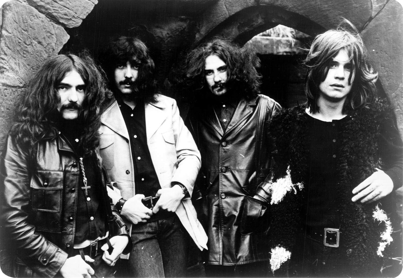 Four men with long hair and moustaches stand in front of a stone arch, wearing leather jackets and serious expressions, in a black and white photo.