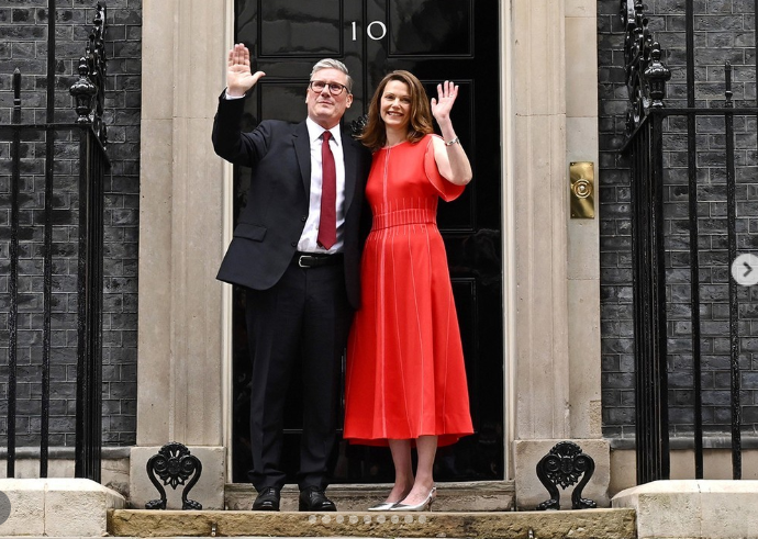 Man in suit and woman in red dress wave outside a black door marked '10', flanked by wrought iron railings and brick walls.