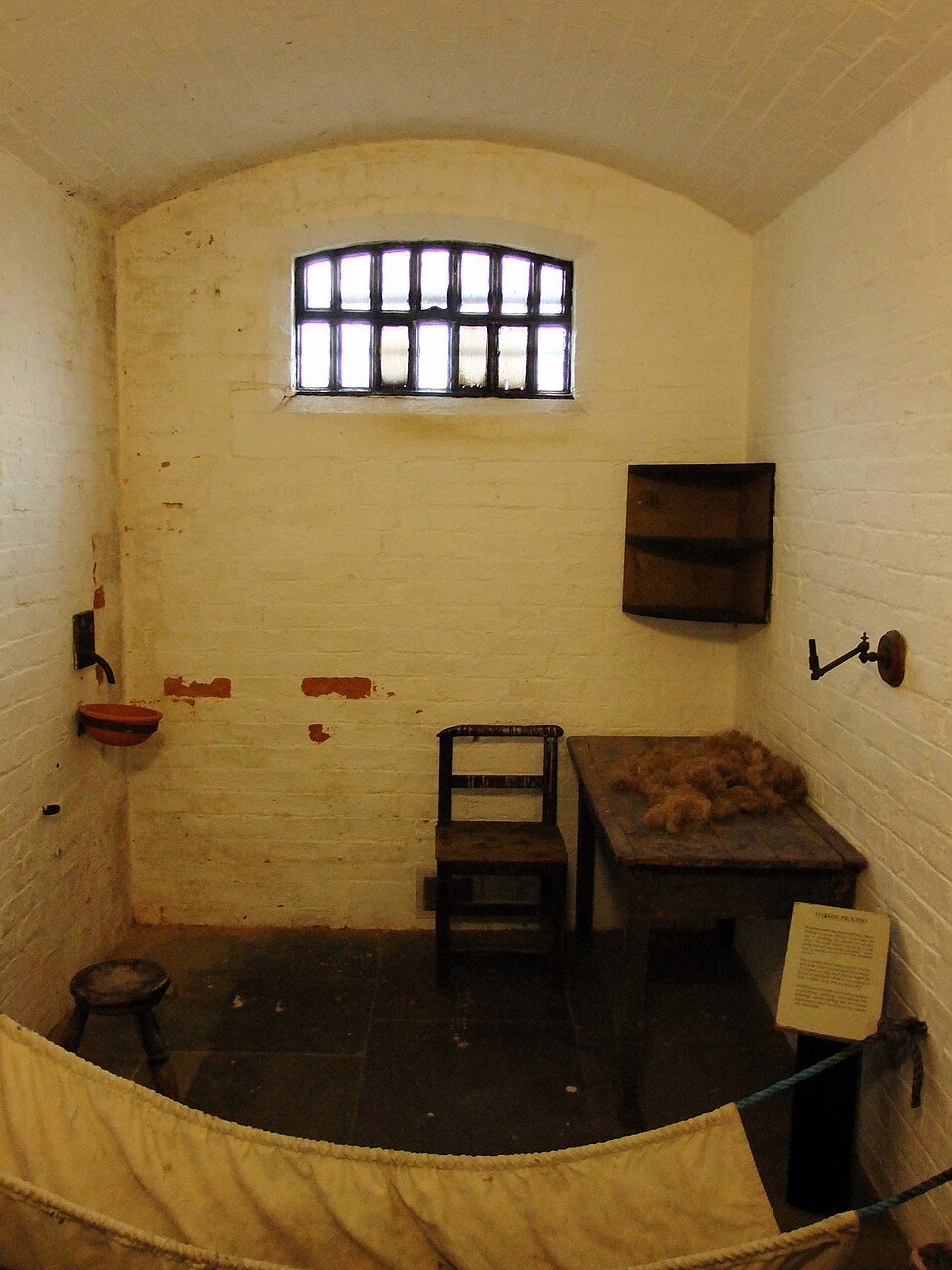 Simple, dimly lit prison cell with a barred window, wooden table, and chair. Sparse furnishings include a hammock and dimly visible shelf.