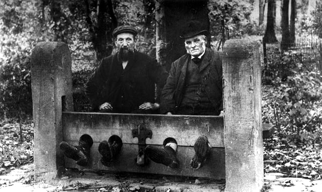 Two elderly men are seated in a wooden stocks in a wooded area, with their feet secured in holes. Both are dressed in dark clothing and wear hats.