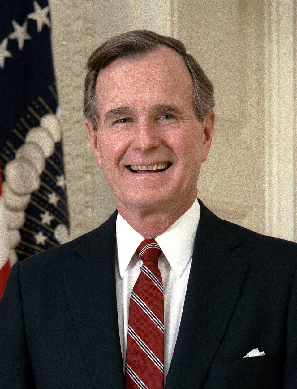 Smiling man in a suit and striped red tie stands indoors with a partial American flag visible in the background.