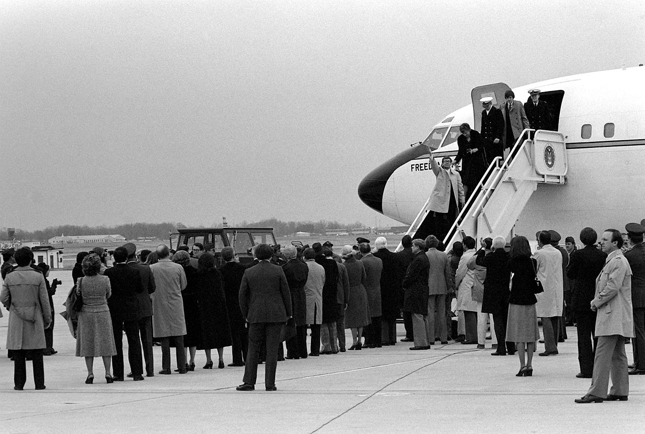 A crowd gathers on an airport tarmac as people disembark from a jet staircase. One person waves while others exit, with spectators observing.