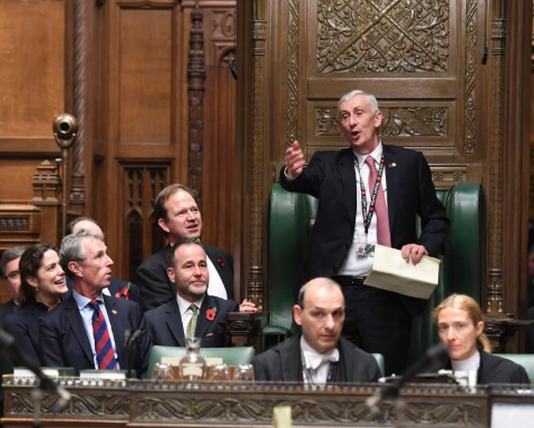A person in a suit stands and speaks animatedly in a wooden-panelled room with several seated individuals, suggesting a formal or parliamentary setting.