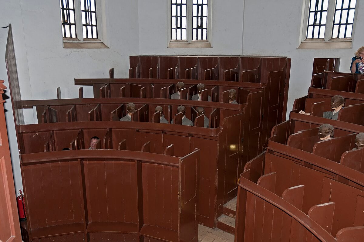 Wooden church pews with individual high partitions, featuring mannequins dressed in period clothing, illuminated by natural light from windows.