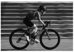 A cyclist in motion rides a racing bicycle along a paved path, wearing a helmet and sports attire, with blurred steps in the background.
