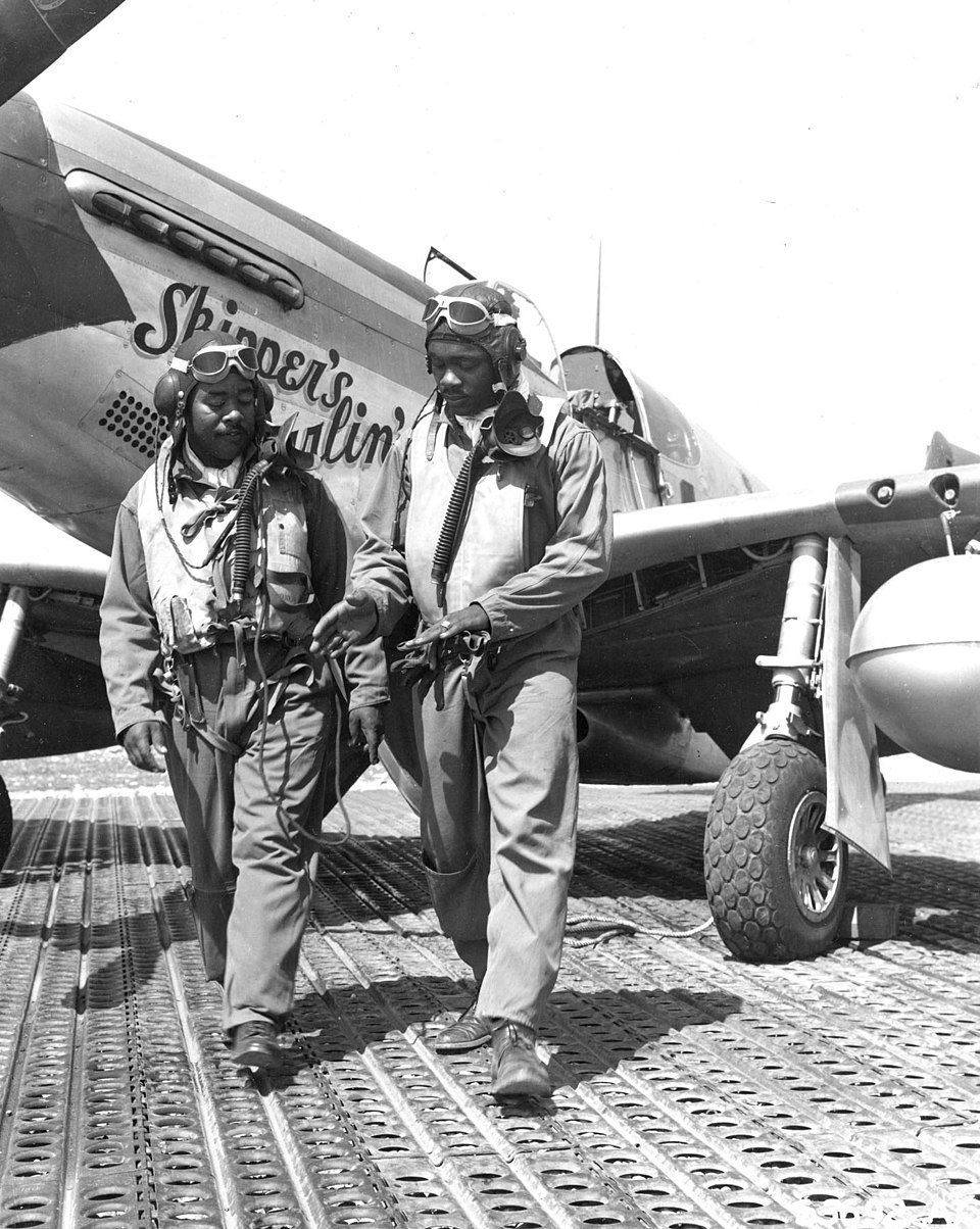 Two pilots in flight gear stand in front of a military aircraft. The plane has "Skipper's Darlin'" painted on its nose.