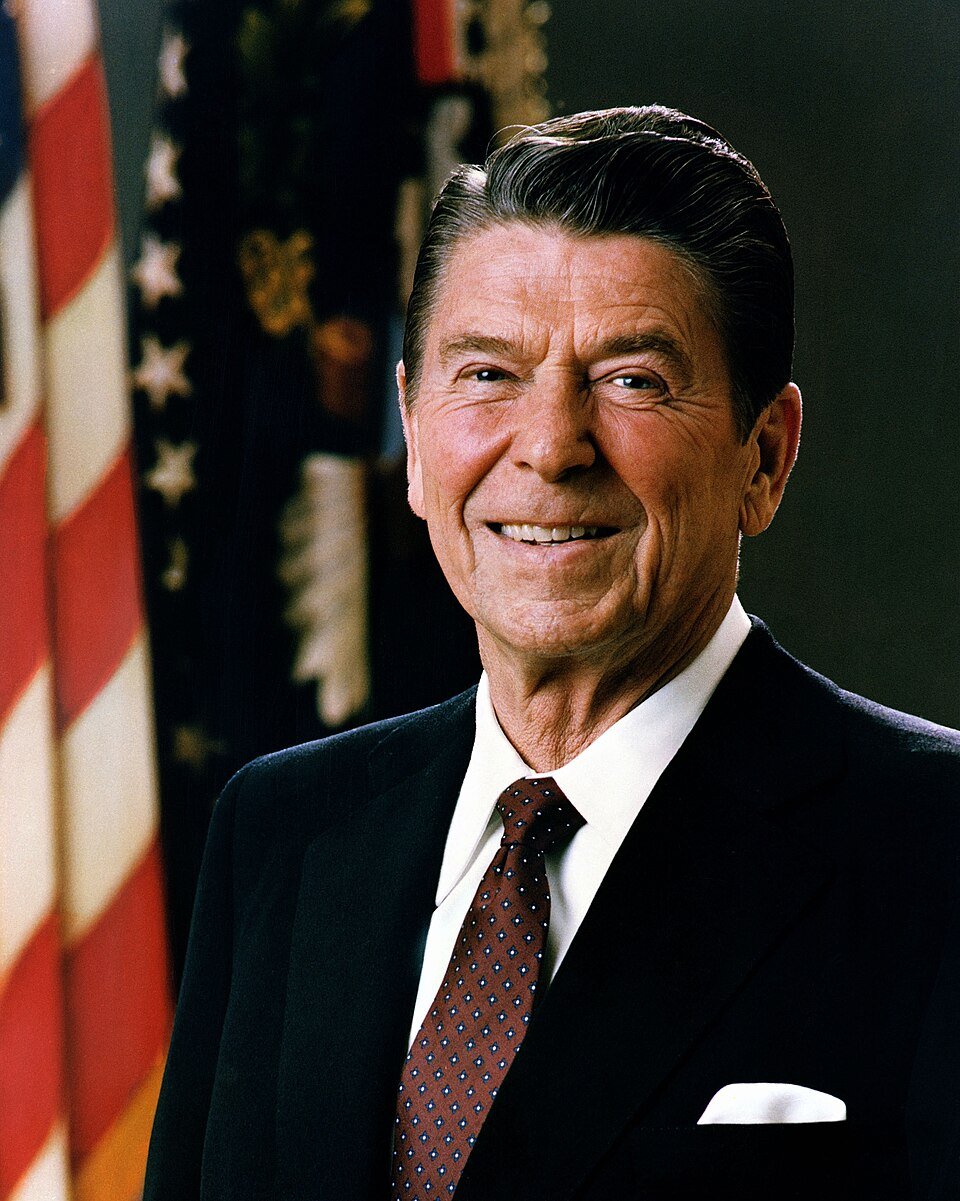 Smiling older man in a suit and tie against a backdrop of the American flag and presidential seal.