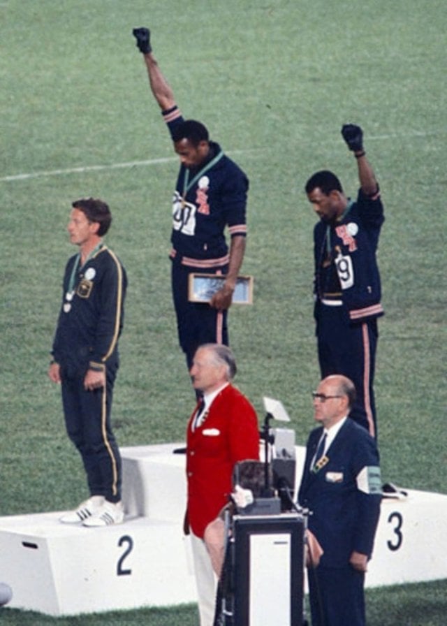 Three athletes on a podium; two wear black gloves and raise fists, while one stands with hands at sides. Officials in suits observe from below.