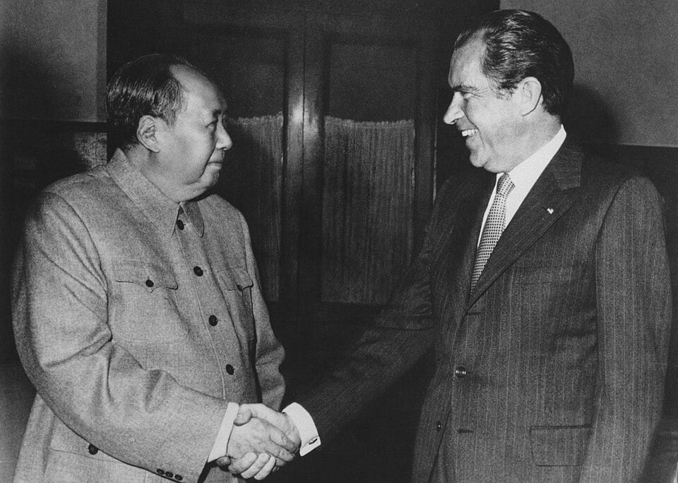Two men, one in a suit and the other in a military-style jacket, shaking hands and smiling inside a dimly lit room.