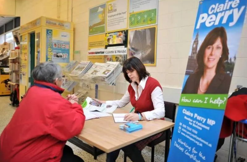 A woman in a red vest holds a meeting at a desk with a man in a red jacket. A large banner displays the name Claire Perry MP.