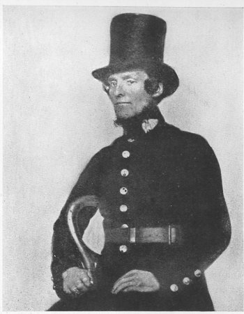A 19th-century man in a formal long coat with buttons and a high top hat, sitting on a chair, with a serious expression and sideburns.