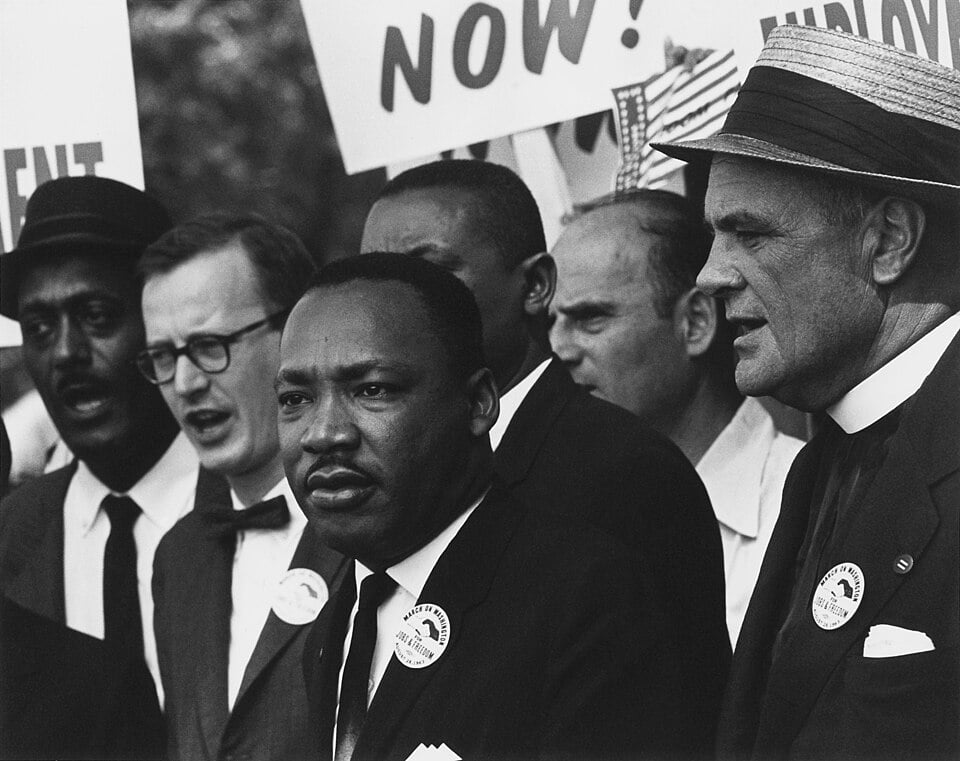 A group of men, some wearing badges, stand together at a protest, holding signs that read "NOW!" and "UNION." One wears a straw hat.