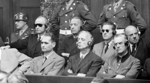 Men seated wearing suits and headphones in a courtroom setting, with military personnel standing behind them, suggestive of a historical trial.