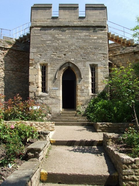 Stone castle entrance with a pointed arch door, flanked by narrow windows. Pathway leads through a garden with plants and flowers to the entrance.