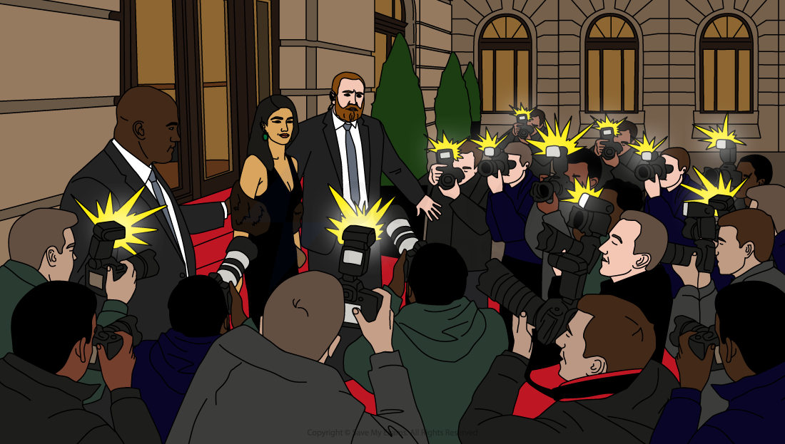 A couple on a red carpet are surrounded by numerous photographers with flashing cameras outside a grand building with arched windows.